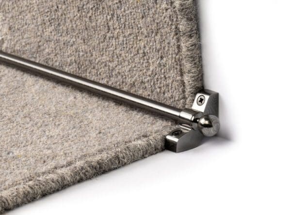 Close-up of a carpet stair rod installation, showing a metallic Stair Rods - Jubilee secured with a bracket to the edge of a grey carpeted step.
