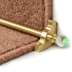 A close-up of a beige carpet held in place on a stair by a gold stair runner rod with a crystal finial.