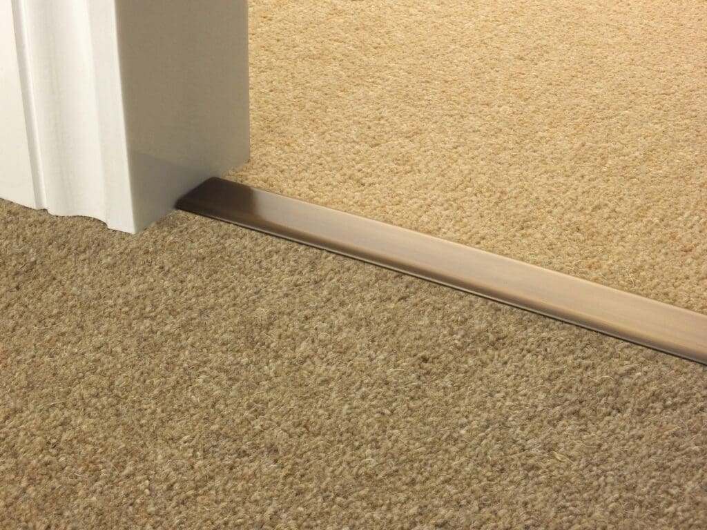 A door partially open showing a Premier Trims - DoubleZ 9 between two different carpeted areas, one slightly darker than the other. The Premier Trims - DoubleZ 9 is metallic and runs beneath the door.