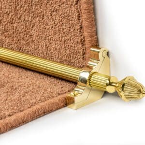 A close-up of a brass stair rod with an ornate finial, installed on a carpeted stair step with a brown carpet runner.