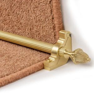 Close-up of a gold-colored stair rod with a decorative finial, securing beige carpet on a staircase.