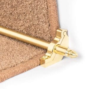 A close-up image of a gold-colored stair rod and bracket set securely positioned at the edge of a beige carpeted stair.