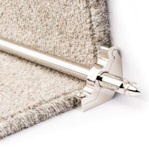 A close-up view of a silver stair rod and bracket holding a beige carpet runner in place on a stair.