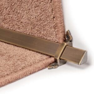 Close-up of a bronze stair rod with brackets securing a beige carpet on a staircase.