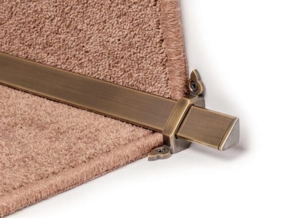 Close-up of a bronze stair rod with brackets securing a beige carpet on a staircase.