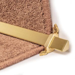 Close-up of a gold stair rod with a decorative bracket securing a brown carpet on a stair step.