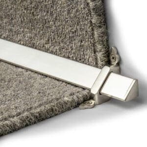 A close-up of a metallic stair rod installed on a carpeted stair, securing the carpet in place with brackets at the ends.