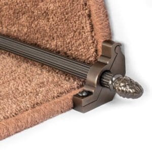 Close-up of a decorative metal stair rod with an ornate end cap, securing a brown carpet runner on a staircase.