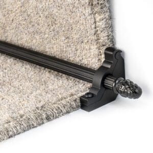 A close-up of a black decorative stair rod securing the edge of a beige carpet on a staircase.