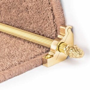 Close-up of a stair carpet holder with a gold-colored rod and ornate finial securing a beige carpet on the edge of a stair step.