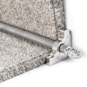 Close-up of a metallic stair rod with decorative finial, securing a light gray carpet on a stair tread.