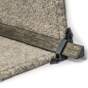 A textured beige carpet stair runner is secured with an angular, dark wooden stair rod and black metal brackets at the end of a staircase.