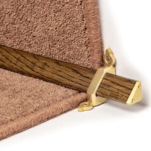 Close-up of a wooden stair rod with brass brackets securing it to the bottom edge of a beige carpeted stair.