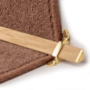 Close-up of a brass stair rod and wooden bracket securing the edge of a brown carpet on a stair step.