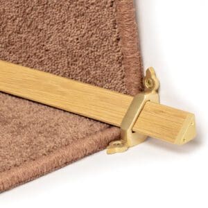 A close-up image of a beige carpet secured by a gold-colored carpet rod and end bracket on a stair edge.