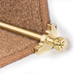 Close-up of a stair rod with a golden finish, secured on a brown carpeted staircase. The rod is held in place by an ornate bracket.