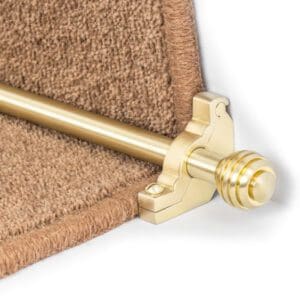 A brass stair rod with decorative end caps secures brown carpet on a stair tread.